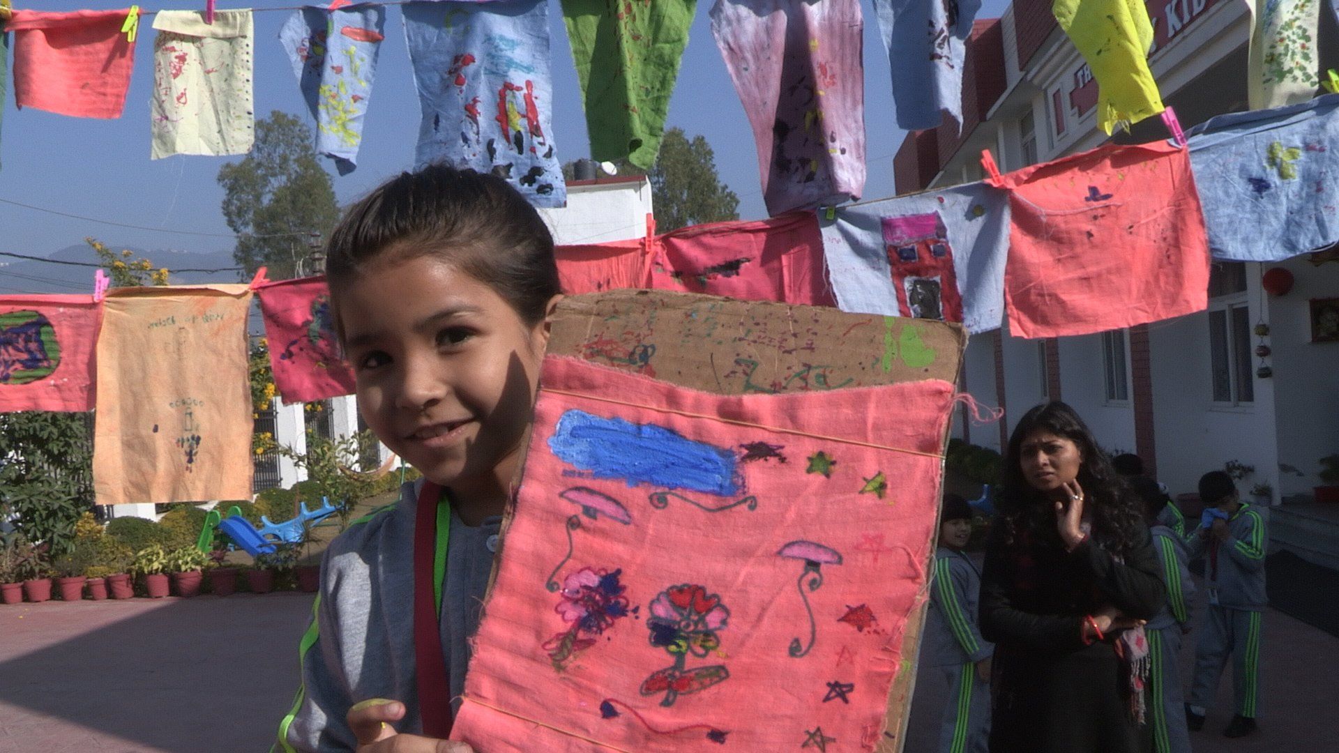 A smiling girl holds up her painted wish flag beneath lines of flags strung across a schoolyard